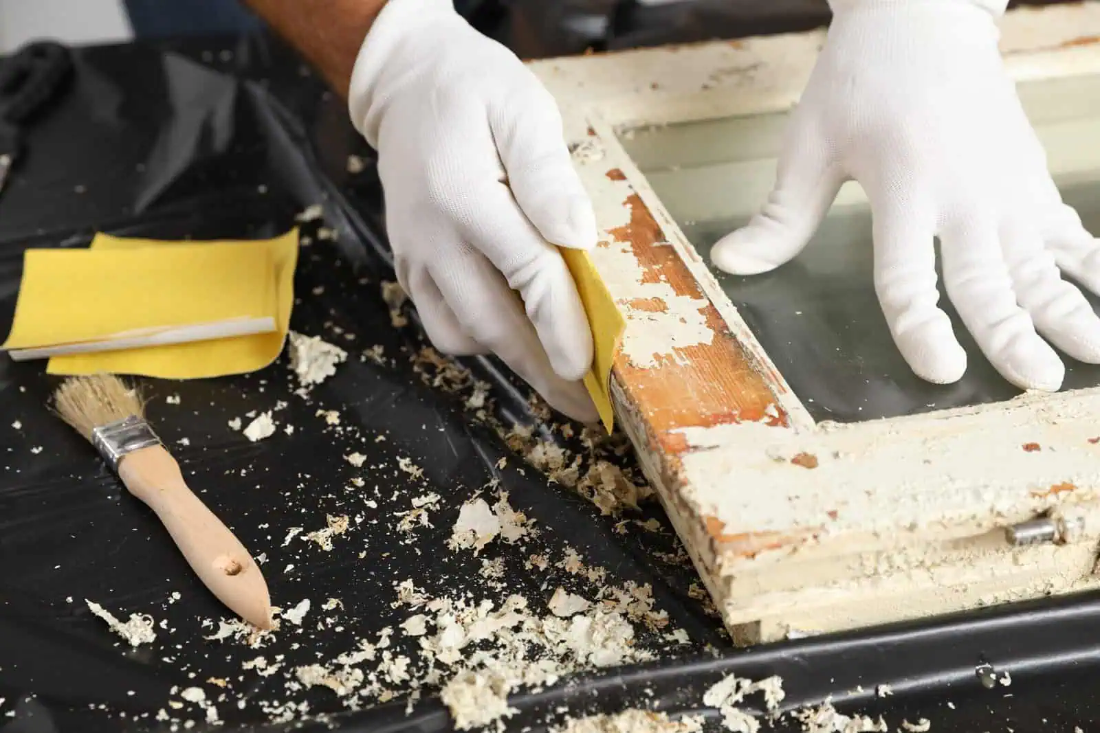 Worker repairing and painting a window frame with tools and gloves.